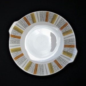 May include: A white ceramic serving dish with a black, yellow, and orange geometric pattern around the rim. The dish has two handles on the sides.