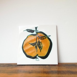 Puede incluir: Azulejo de cerámica cuadrado con una ilustración dibujada a mano de una manzana cortada. La manzana es naranja con contornos y detalles negros, sobre un fondo blanco. El azulejo se muestra sobre una superficie de madera.