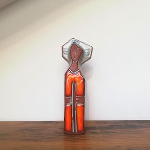 May include: A ceramic figurine with a stylized human form. The figure has a red-orange body with vertical white lines and a brown face. The head is topped with a silver-coloured halo. The figurine is standing on a wooden surface.