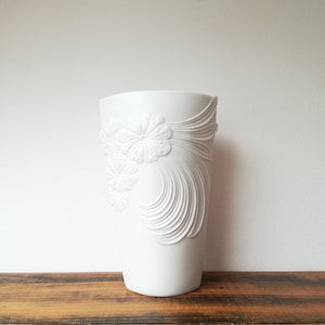 May include: White ceramic vase with a raised floral design. The vase has a cylindrical shape and a smooth, white finish.