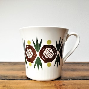 May include: White ceramic mug with a brown, green, and yellow geometric pattern. The pattern features stylized flowers and leaves.
