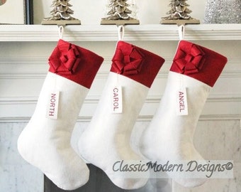 May include: Three white Christmas stockings with red velvet tops and bows, hanging from a white mantel with silver Christmas tree ornaments. The stockings are personalized with the names "North", "Carol", and "Angel".