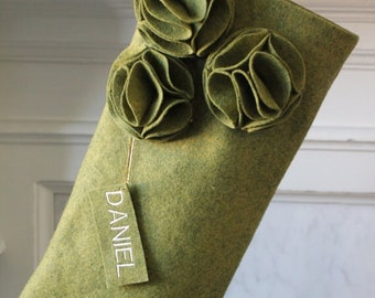 May include: Olive green felt Christmas stocking with three decorative flower accents. A tag with the name "DANIEL" hangs from the stocking. The words "modern felt desings" are printed on the stocking.