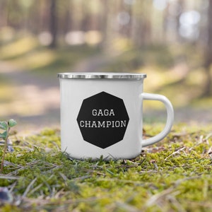 Può includere: Tazza da campeggio in smalto bianco con una forma ottagonale nera che dice "GAGA CHAMPION" in testo bianco. La tazza è appoggiata su un letto di muschio verde.