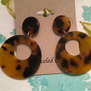 May include: A pair of brown and black tortoise shell earrings with a gold accent. The earrings are made of two circles, one smaller and one larger. The larger circle has a hole in the center.