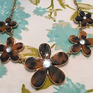 May include: A gold chain necklace with five brown and gold flower pendants. Each flower has a clear rhinestone in the center.