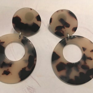 May include: A pair of large, round, dangle earrings with a tortoiseshell pattern. The earrings are made of a cream-colored material with brown and black swirls.