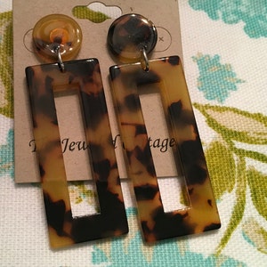 May include: A pair of brown and black rectangular earrings with a smaller rectangle cut out in the center. The earrings have a tortoiseshell pattern.