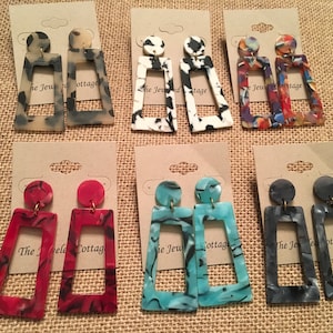 May include: Assortment of 10 pairs of colorful rectangular statement earrings with a variety of marbled patterns. The earrings are made of resin and have a gold-tone hook closure. The earrings are displayed on brown card stock with the text "The Jeweled Cottage" printed on each card.