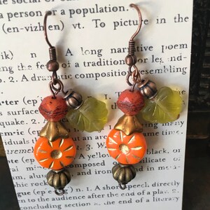 May include: A pair of earrings with orange, green, and brown glass beads. The earrings are made with copper wire and have a hook closure.