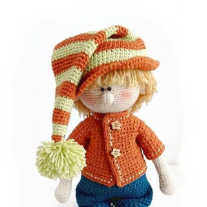 Puede incluir: Una mu&ntilde;eca de crochet con pelo rubio que lleva un gorro a rayas naranja y verde, un jersey naranja con botones, pantalones azules y zapatos marrones.