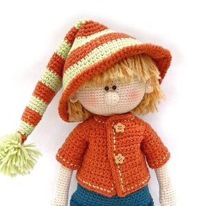 Puede incluir: Una mu&ntilde;eca de crochet con un sombrero a rayas, un su&eacute;ter naranja, pantalones azules y zapatos marrones. La mu&ntilde;eca tiene el pelo rubio y una cara sonriente.
