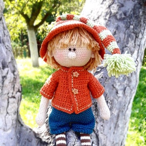 Puede incluir: Una mu&ntilde;eca de crochet con un gorro a rayas rojas y blancas, un su&eacute;ter naranja, pantalones azules y piernas a rayas marrones y blancas. La mu&ntilde;eca est&aacute; sentada en un tronco de &aacute;rbol.