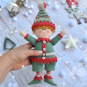May include: A green crocheted elf doll with a red and white striped hat, red and white striped socks, and a red and white striped belt. The elf has a green and white striped hat with a white pom-pom on top. The elf has a red and white striped belt with a white buckle. The elf has a red and white striped hat with a white pom-pom on top.