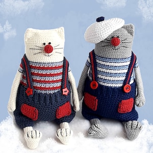 May include: Two crocheted cat toys wearing sailor outfits. One cat is white with a red nose and the other is gray with a white sailor hat. Both cats are wearing blue overalls with red accents and white striped shirts.