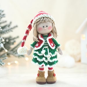 Op de afbeelding: Een handgemaakte gehaakte pop in een feestelijke kerstoutfit. De pop heeft blond haar en roze wangen, draagt een groene jurk met witte rand, een rood-witte jas en een bijpassende hoed met een lange, gestreepte staart. De pop draagt bruine laarzen.