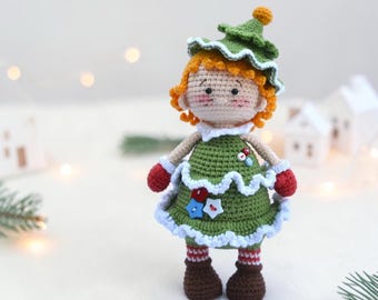 May include: A handmade crochet doll in a Christmas tree costume. The doll has orange curly hair, a green tree-shaped hat, and a green dress with white trim. It has red mittens, striped socks, and brown boots. The doll is decorated with blue and red star-shaped embellishments.