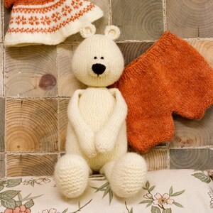 Puede incluir: Un oso de peluche blanco de punto que lleva un vestido de punto marr&oacute;n y pantalones cortos de punto marr&oacute;n. El oso est&aacute; sentado sobre una superficie con estampado floral.
