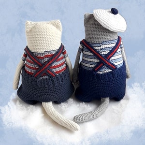 May include: Two crocheted cat toys, one white and one gray, wearing blue overalls with red and white striped shirts. The white cat has a white tail and the gray cat has a gray tail. Both cats are sitting on a white fluffy surface.