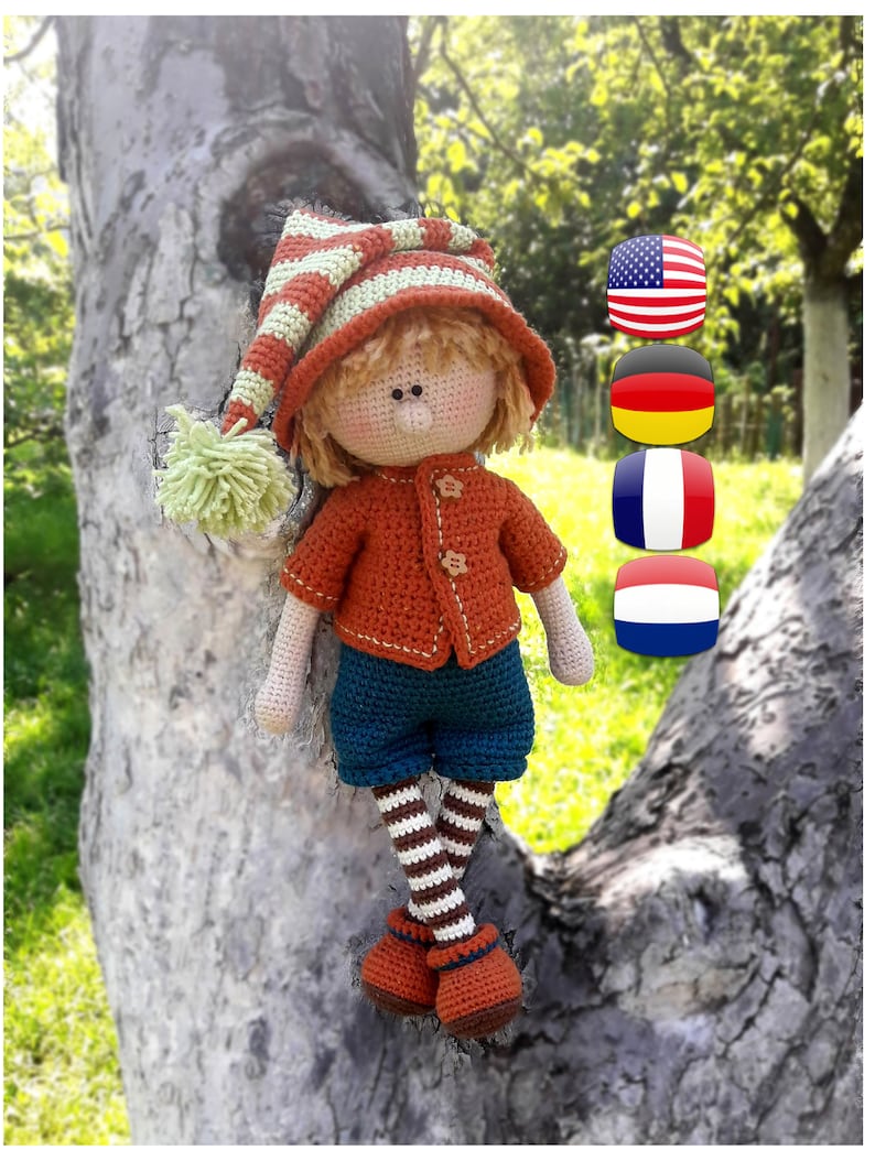 Puede incluir: Una mu&ntilde;eca de crochet con un sombrero a rayas, un su&eacute;ter naranja, pantalones cortos azules y medias a rayas. La mu&ntilde;eca est&aacute; sentada en un tronco de &aacute;rbol con un fondo verde. La imagen tambi&eacute;n incluye banderas de los Estados Unidos, Alemania, Francia y los Pa&iacute;ses Bajos.