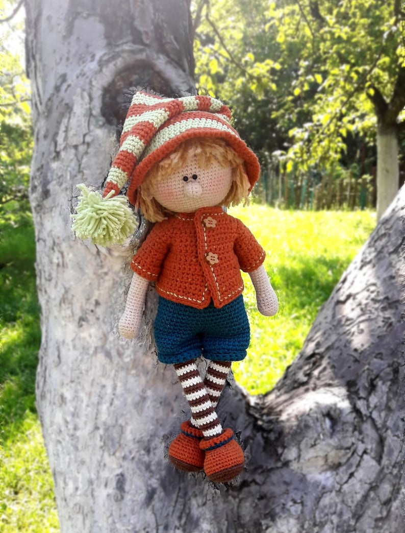 Puede incluir: Una mu&ntilde;eca de crochet con un gorro a rayas, un su&eacute;ter naranja, pantalones azules y medias a rayas. La mu&ntilde;eca est&aacute; sentada en un tronco de &aacute;rbol.