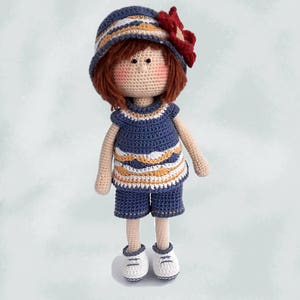 May include: A crocheted doll with brown hair, wearing a blue and white striped hat with a red flower, a blue and white striped dress, blue shorts, and white sneakers.