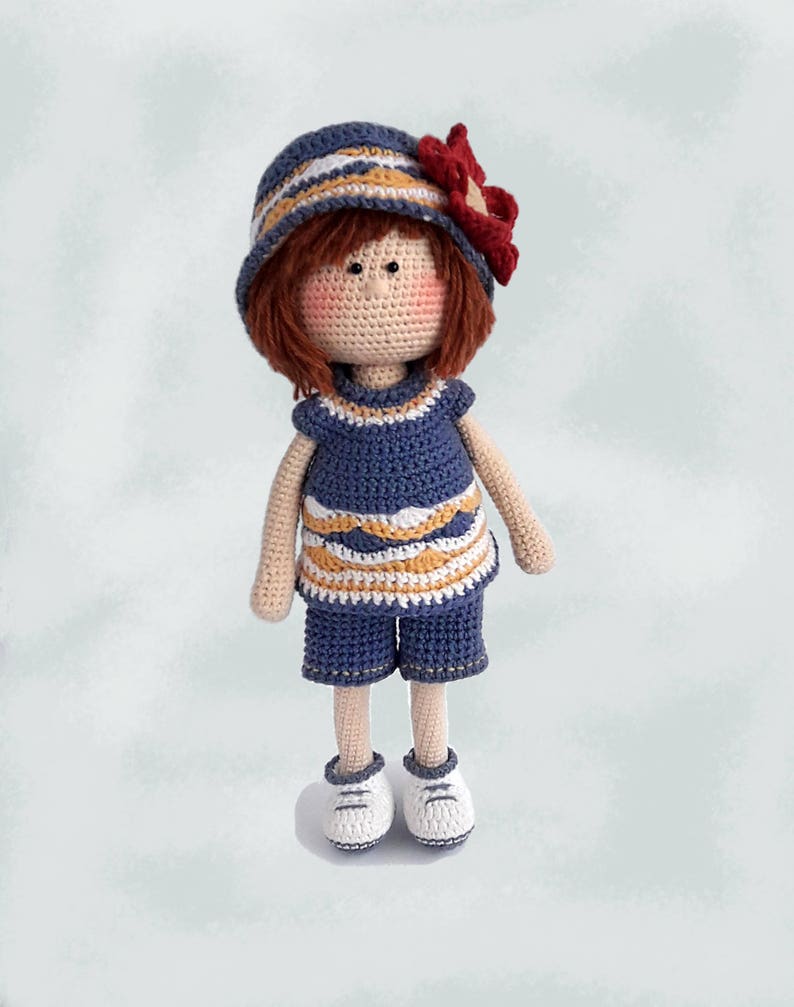 May include: A crocheted doll with brown hair, wearing a blue and white striped hat with a red flower, a blue and white striped dress, blue shorts, and white sneakers.