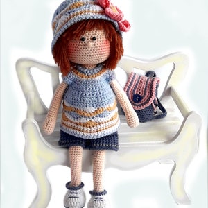May include: A crocheted doll wearing a blue and white striped dress, a blue hat with a pink flower, and blue shoes. The doll is sitting on a white wooden bench with a pink and blue striped backpack.