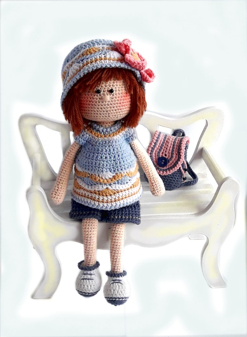 May include: A crocheted doll wearing a blue and white striped dress, a blue hat with a pink flower, and blue shoes. The doll is sitting on a white wooden bench with a pink and blue striped backpack.