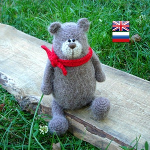 May include: A gray crocheted teddy bear wearing a red scarf sits on a wooden surface. The bear has black eyes and a brown nose. The background is a green grassy area.