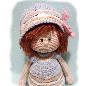 May include: A crocheted doll wearing a blue and white striped dress and a blue and white striped hat with a pink flower. The doll has brown hair and brown eyes.