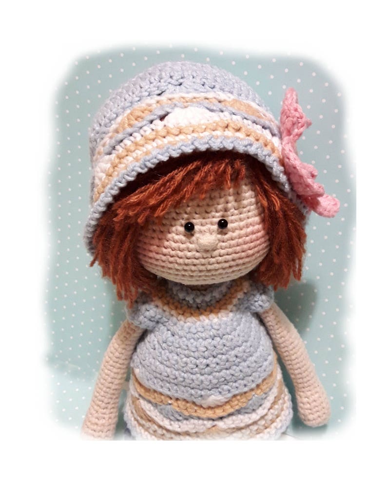 May include: A crocheted doll wearing a blue and white striped dress and a blue and white striped hat with a pink flower. The doll has brown hair and brown eyes.