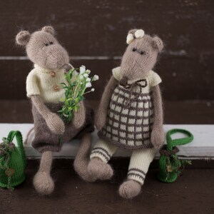 May include: Two knitted plush mice wearing clothes and holding a bouquet of white flowers. The mouse on the left is wearing a yellow shirt and brown trousers. The mouse on the right is wearing a plaid dress and white tights. Both mice are holding small green bags.