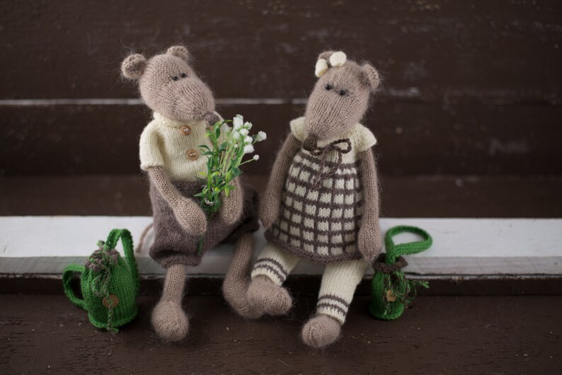 May include: Two knitted plush mice wearing clothes and holding a bouquet of white flowers. The mouse on the left is wearing a yellow shirt and brown trousers. The mouse on the right is wearing a plaid dress and white tights. Both mice are holding small green bags.