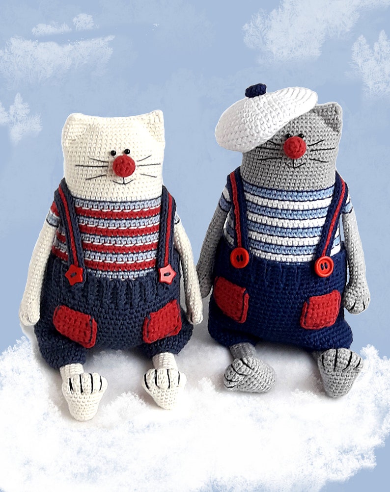 May include: Two crocheted cat toys wearing sailor outfits. One cat is white with a red nose and the other is gray with a white sailor hat. Both cats are wearing blue overalls with red accents and white striped shirts.