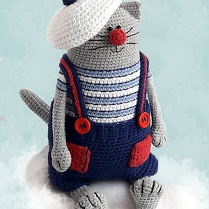May include: A gray crocheted cat wearing a white crocheted beret, a blue and white striped shirt, and blue overalls with red accents. The cat is sitting on a white fluffy surface.