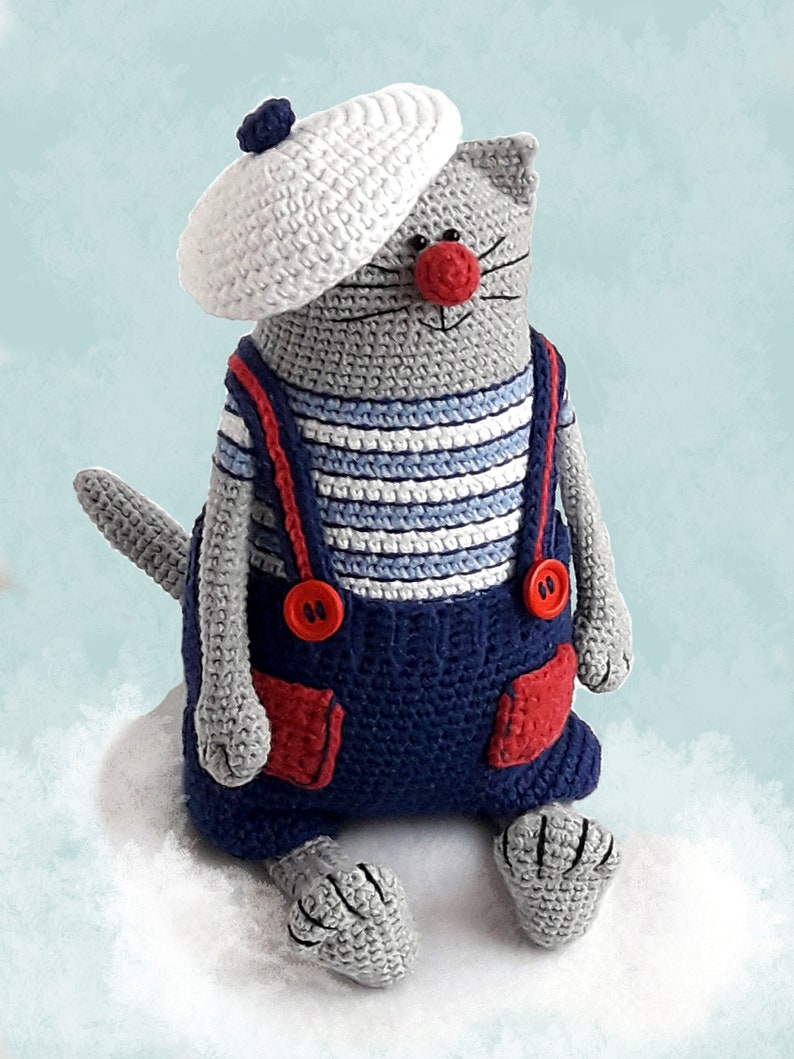 May include: A gray crocheted cat wearing a white crocheted beret, a blue and white striped shirt, and blue overalls with red accents. The cat is sitting on a white fluffy surface.
