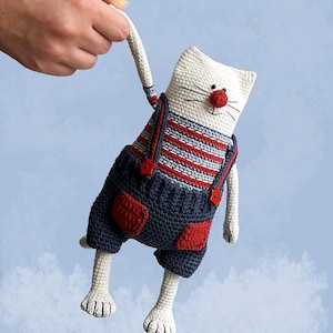 May include: White crocheted cat toy wearing blue overalls with red accents and a red nose. The cat is holding a red button in its mouth.