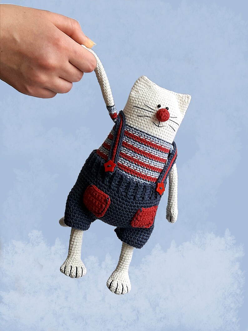 May include: White crocheted cat toy wearing blue overalls with red accents and a red nose. The cat is holding a red button in its mouth.