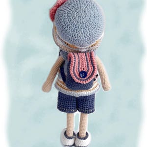 May include: A crocheted doll wearing a blue and white striped shirt, blue shorts, a blue hat with a pink bow, and a pink backpack with a blue button. The doll has brown yarn hair and is standing on a light blue background.