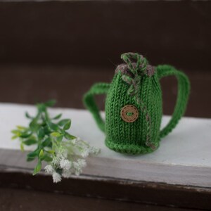 May include: A small green knitted backpack with a brown button and brown straps. The backpack is sitting on a white surface with a small green plant in the background.