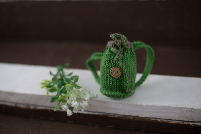 May include: A small green knitted backpack with a brown button and brown straps. The backpack is sitting on a white surface with a small green plant in the background.