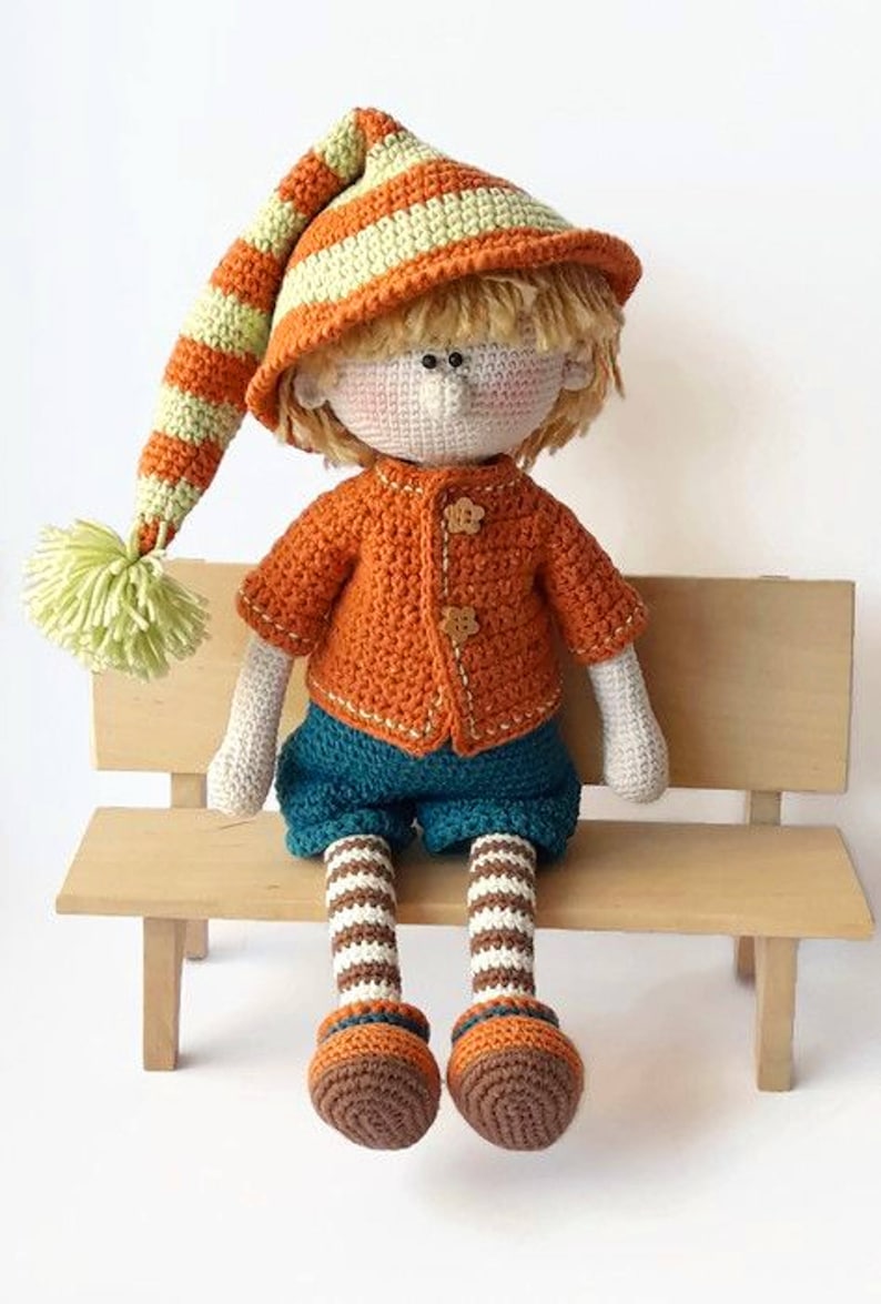 Puede incluir: Una mu&ntilde;eca de crochet con un sombrero a rayas, un su&eacute;ter naranja, pantalones azules y calcetines a rayas. La mu&ntilde;eca est&aacute; sentada en un banco de madera.