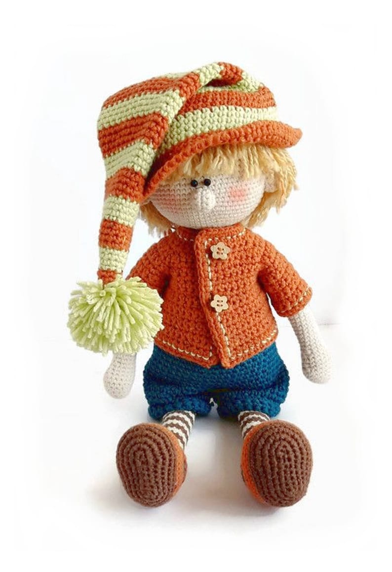Puede incluir: Una mu&ntilde;eca de crochet con pelo rubio que lleva un gorro a rayas naranja y verde, un jersey naranja con botones, pantalones azules y zapatos marrones.
