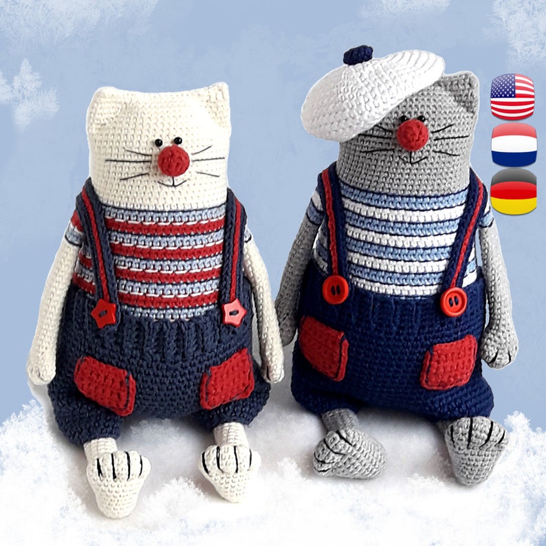 May include: Two crocheted cat toys wearing blue overalls with red accents and striped shirts. One cat is white with a red nose and the other is gray with a white sailor hat.