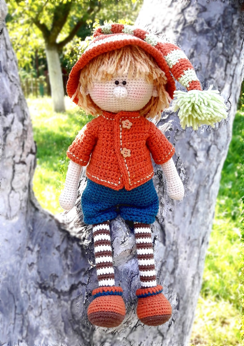 Puede incluir: Una mu&ntilde;eca de crochet con un gorro a rayas rojas y blancas, un su&eacute;ter naranja, pantalones azules y piernas a rayas marrones y blancas. La mu&ntilde;eca est&aacute; sentada en un tronco de &aacute;rbol.