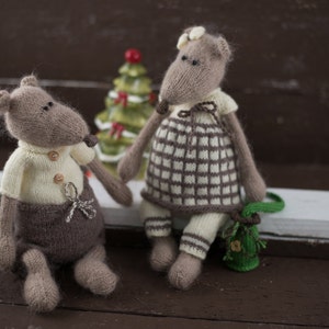May include: Two knitted plush toys shaped like mice. The mouse on the left is wearing a white sweater and brown trousers. The mouse on the right is wearing a white and brown checked dress. Both mice have brown fur and black eyes.
