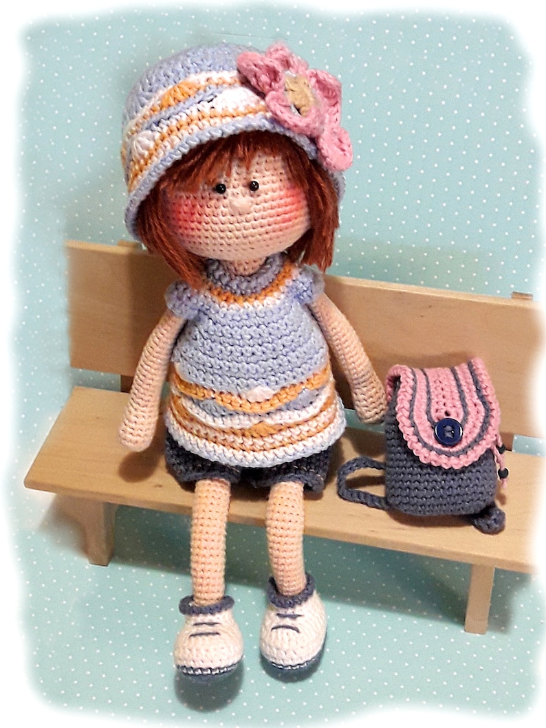 May include: A crocheted doll wearing a blue and white striped hat with a pink flower, a blue and orange striped dress, blue shorts, and white shoes. The doll is sitting on a wooden bench with a gray and pink backpack.