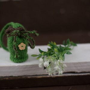 May include: A small green knitted pouch with a brown ribbon and a wooden flower button. The pouch is sitting on a white surface with a sprig of white flowers.
