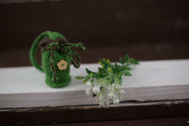 May include: A small green knitted pouch with a brown ribbon and a wooden flower button. The pouch is sitting on a white surface with a sprig of white flowers.